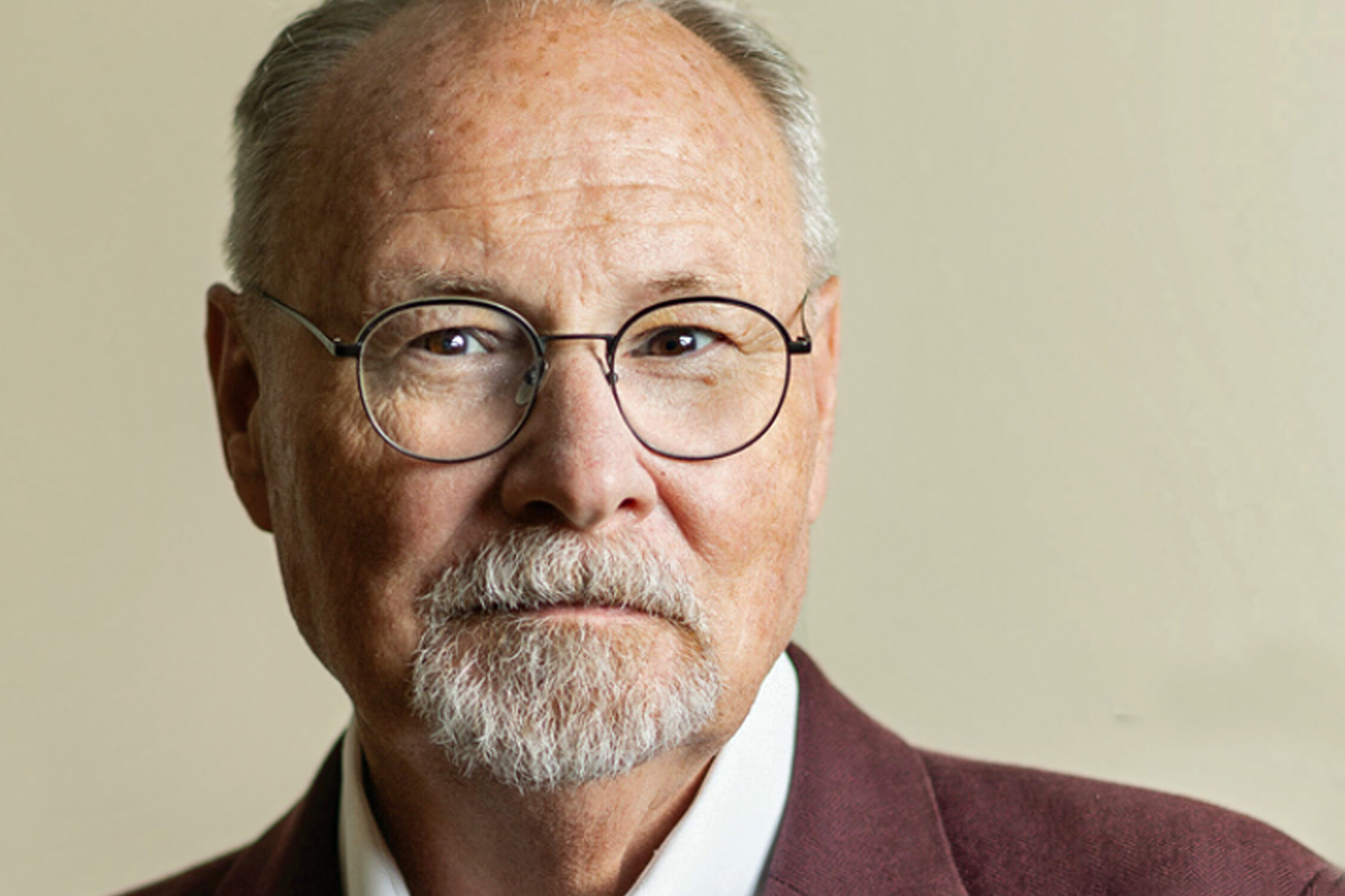An older man with a gray beard and glasses wears a maroon blazer and white shirt. He looks directly at the camera against a neutral background.
