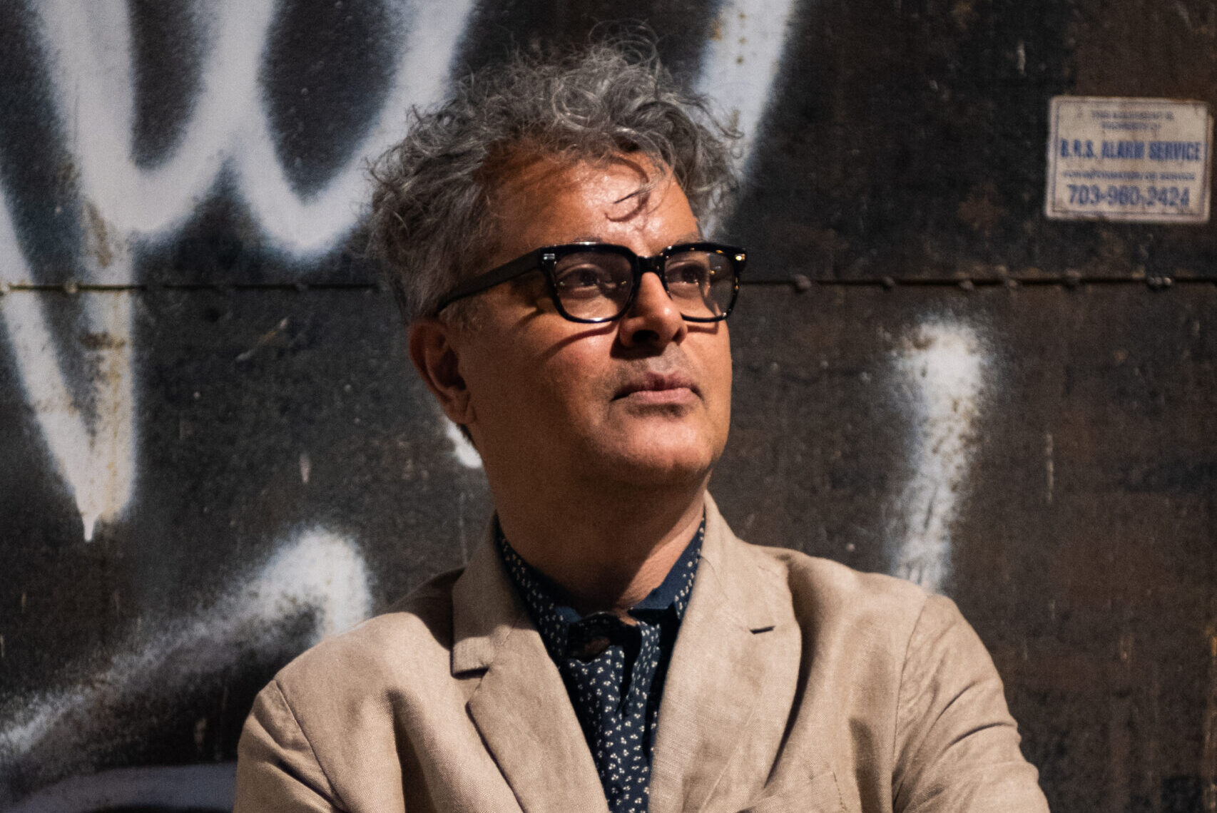 A man with curly gray hair and glasses stands with his arms crossed, wearing a beige suit and navy polka dot tie. He is looking up slightly, in front of a dark wall with graffiti.