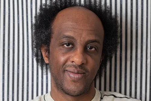 A person with short, curly hair and a slight smile, wearing a light-colored sweater with dark horizontal stripes. They are standing against a vertically striped background.