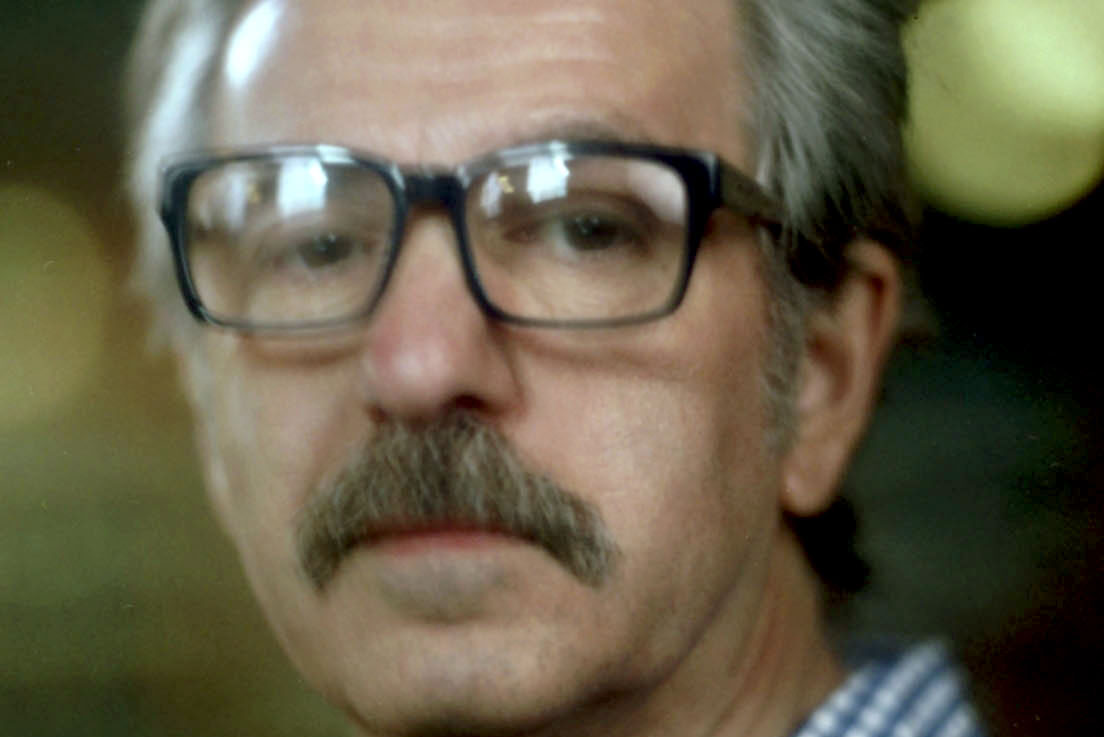 A person with gray hair and a mustache is wearing oversized glasses and a checkered shirt. They are looking directly at the camera. The background is softly blurred with warm lighting.