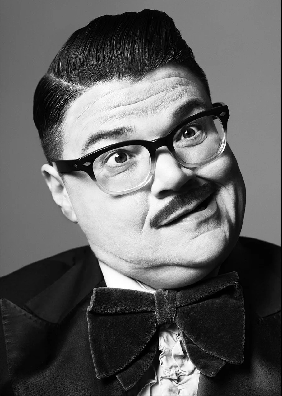 A person with slicked-back hair, thick black glasses, and a small mustache wears a large bow tie and suit, posing with a surprised, wide-eyed expression against a plain background.