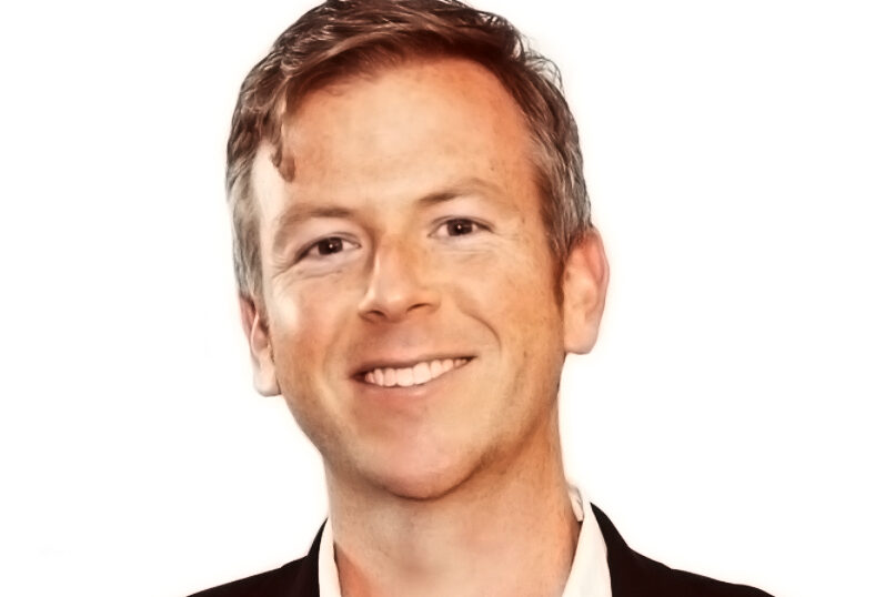 Smiling man with short light brown hair wearing a black suit and white shirt against a plain white background.