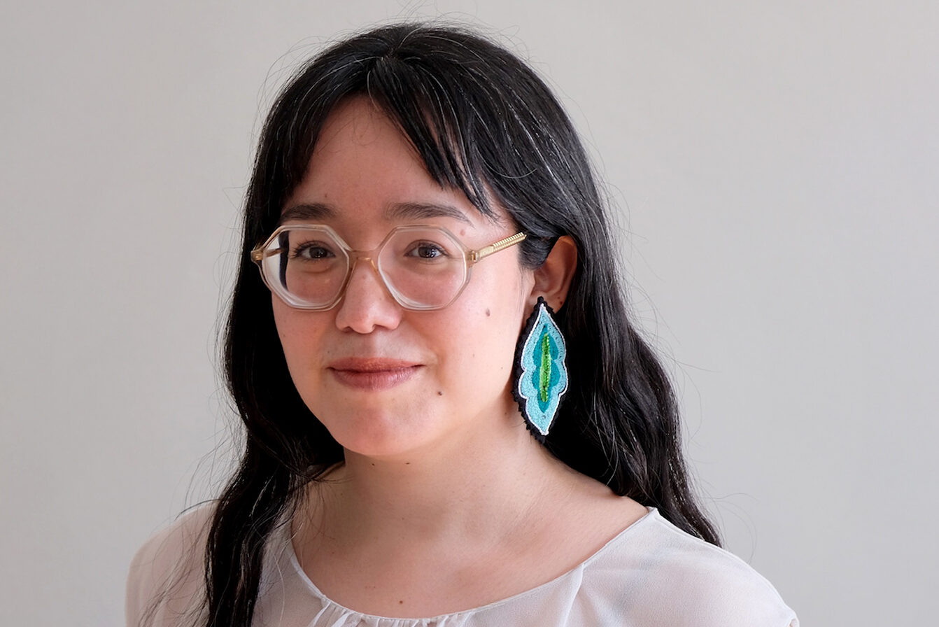A person with long dark hair and glasses is wearing a sheer white top and large blue geometric earrings. They have a tattoo on their left arm and are standing against a plain background, smiling gently.