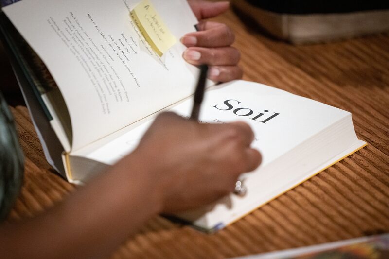 A person is holding a book open, with a hand steadying the page titled Soil. The other hand is writing with a pen. The book lies on a wooden table, and a sticky note is visible on the open pages.