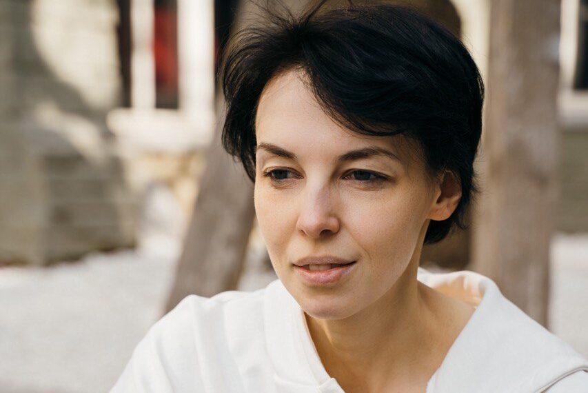 Person with short dark hair sits outdoors, wearing a white sweater and dark pants, with hands clasped around their knees. They appear thoughtful, looking slightly to the side, with a light, blurred background.