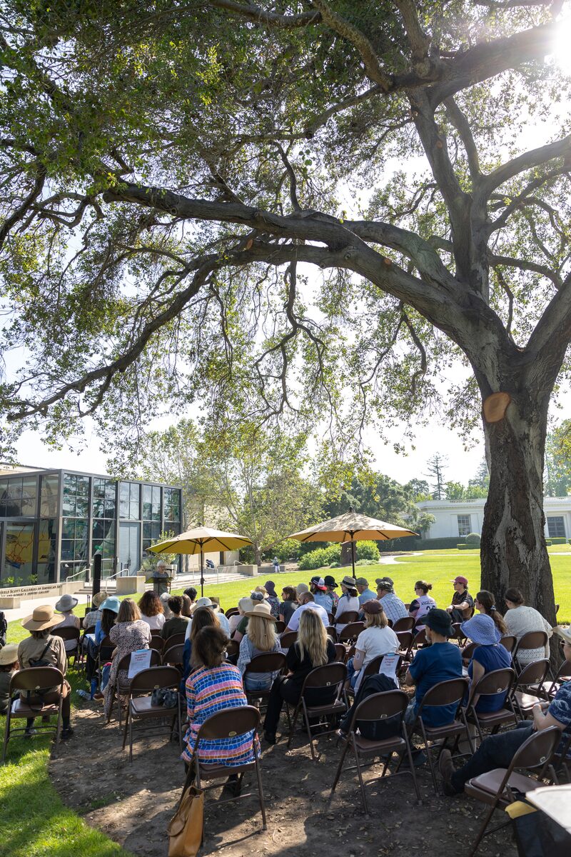 A group of people sitting on chairs outdoors under a large tree, facing a speaker near a modern building with glass windows. Sunlight filters through the leaves, and some attendees have umbrellas for shade.