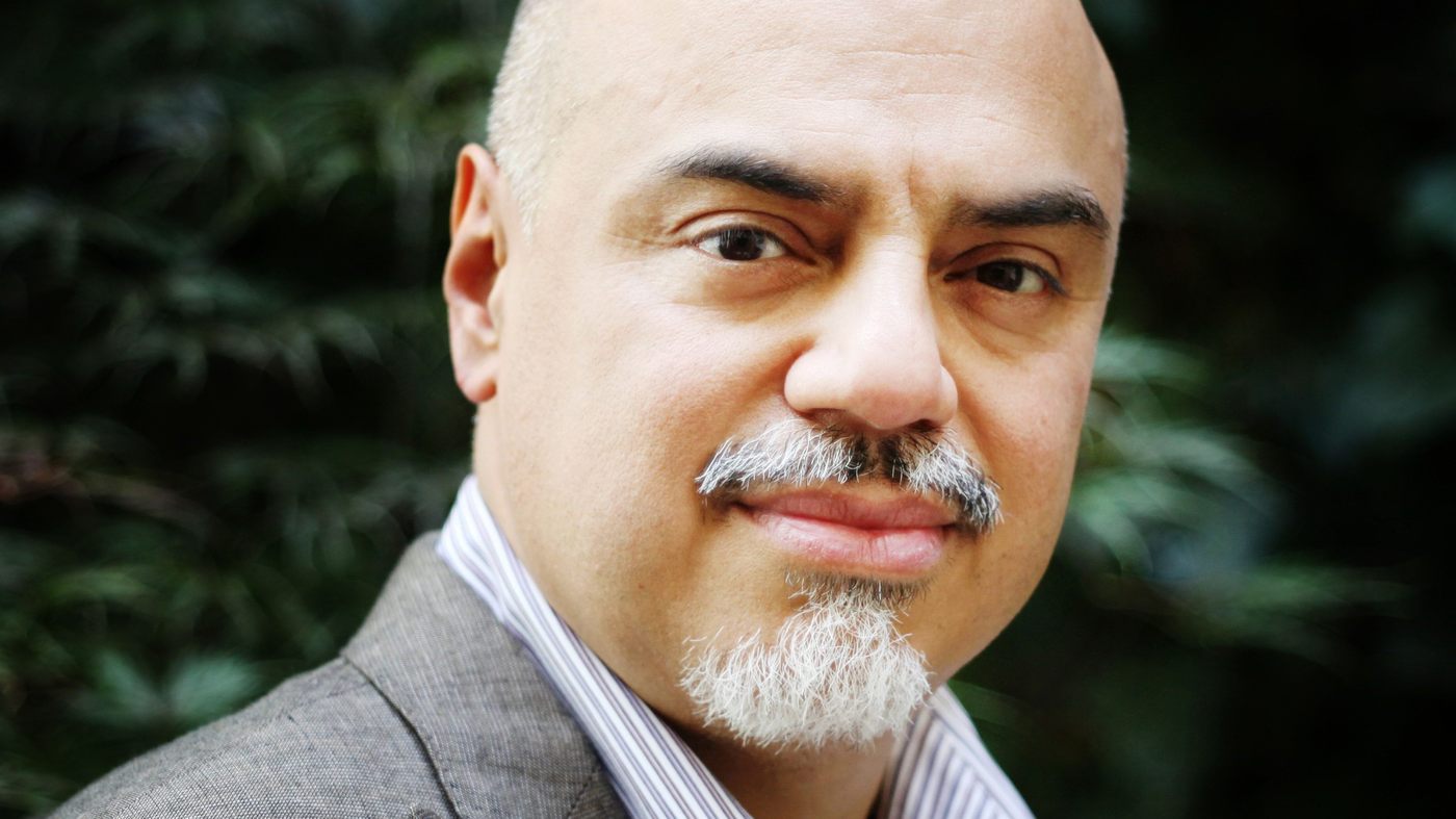 A person with a bald head and goatee is wearing a gray suit with a striped shirt. They are looking at the camera with a slight smile. The background is blurred greenery.