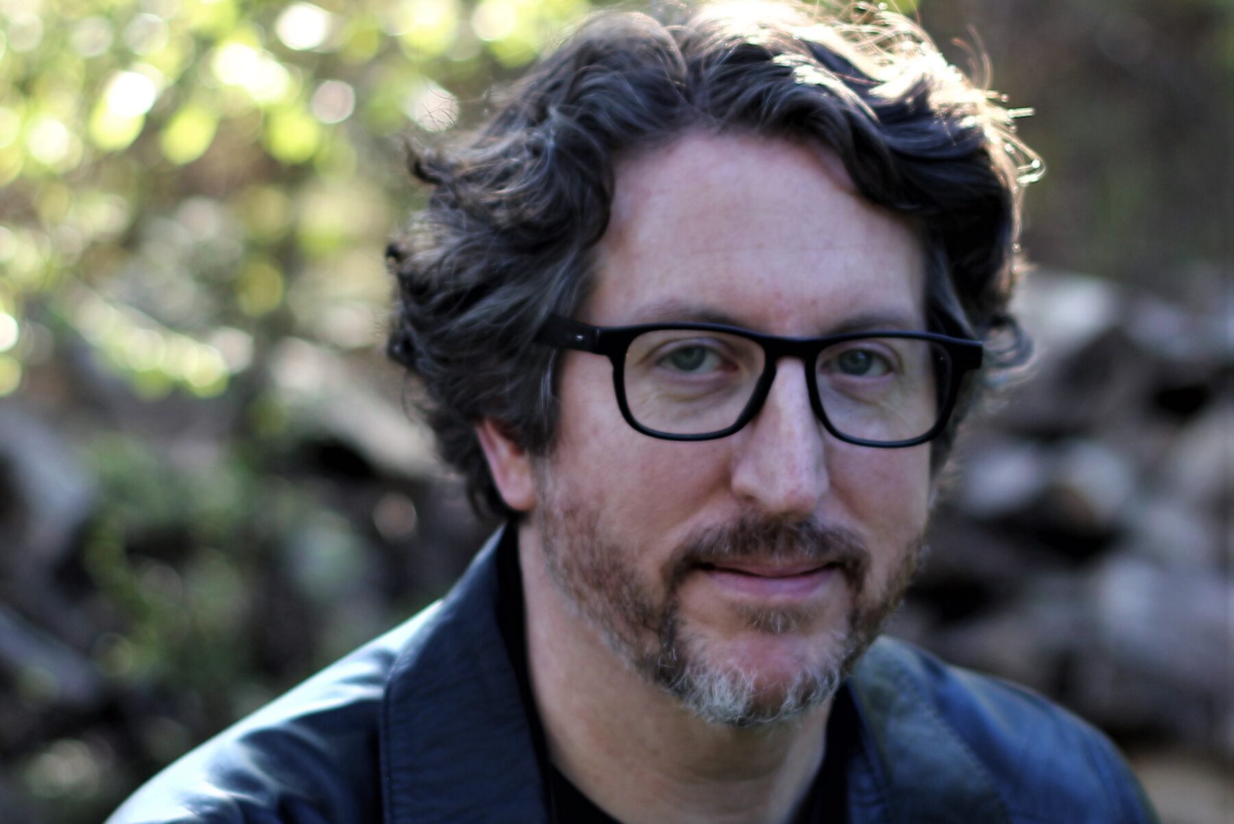 Man with wavy hair and glasses wearing a dark leather jacket, seated outdoors. The background is softly focused, showing greenery and logs, suggesting a natural setting.