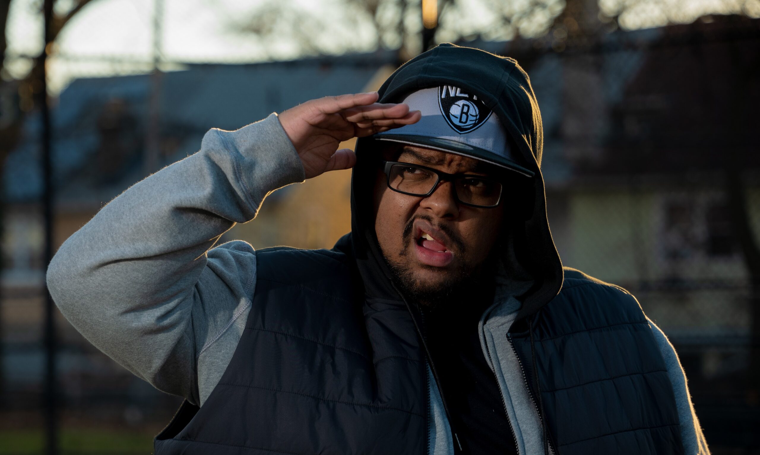 A man wearing glasses, a hoodie, and a Brooklyn Nets cap salutes with his right hand while making a playful facial expression outdoors, with blurred houses and trees in the background.