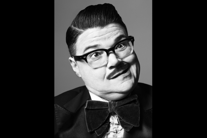 Black-and-white portrait of a person with slicked-back hair, dark-rimmed glasses, a mustache, and a large bow tie, tilting their head and making a quirky facial expression.